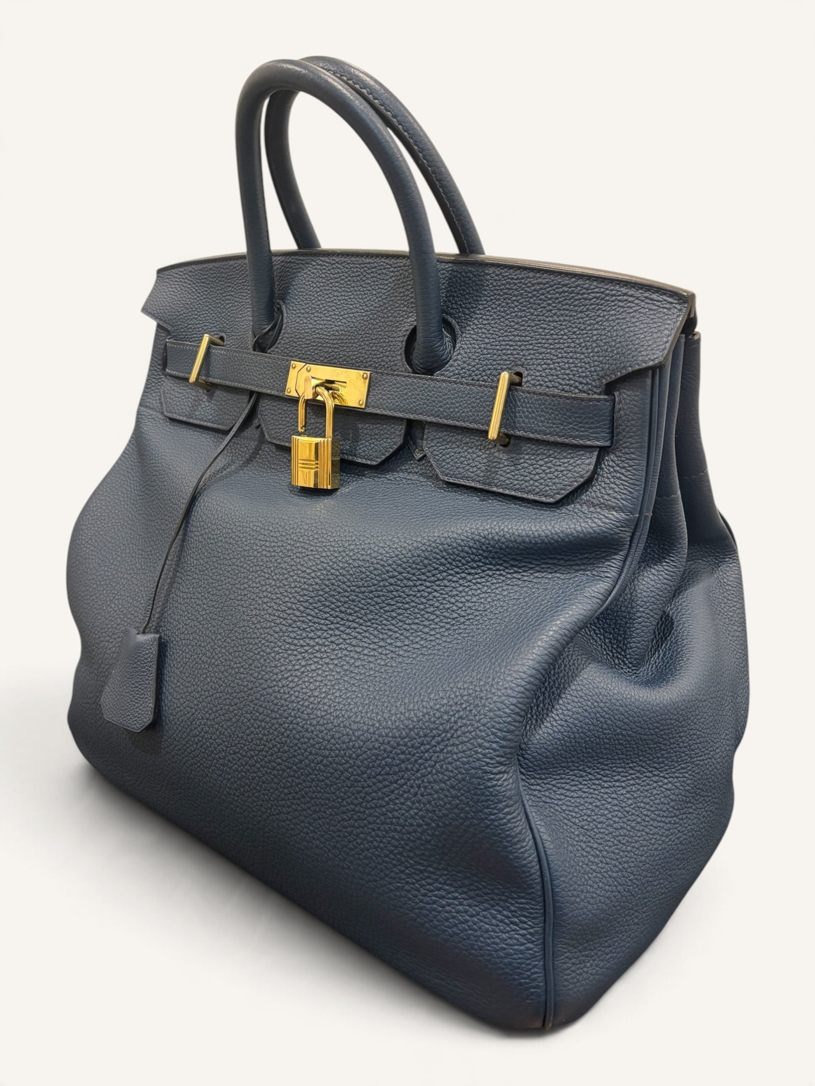 # Hermès HAC 40 Bleu de Prusse en cuir Togo, finitions Gold – 2015 Ce **HAC 40 Bleu de Prusse** en cuir Togo date de **2015**. Il se distingue par son magnifique bleu profond, associé à des **finitions Gold** qui apportent un contraste chaleureux. Présenté en **très bon état général**, cet exemplaire possède uniquement de légères traces d'utilisation au niveau des coins. Son format 40 et sa silhouette caractéristique lui confèrent une présence remarquable. **Dimensions : format 40 cm – dimensions complètes à compléter** **Matière : cuir Togo** **Couleur : Bleu de Prusse** **Finitions : Gold** **Année : 2015** **État : très bon état général, légères traces aux coins** Cette configuration associe l'élégance d'une couleur profonde à la texture du cuir Togo. Ainsi, le sac conserve une esthétique intemporelle tout en se distinguant des coloris plus traditionnels. --- ## HAC 40 Bleu de Prusse : une couleur profonde et élégante Le **Bleu de Prusse** fait partie des tonalités sombres particulièrement intéressantes de la palette Hermès. Sa profondeur lui donne une élégance proche de celle d'un bleu nuit. Cependant, ses nuances peuvent évoluer selon la lumière. Sous un éclairage naturel, le cuir peut révéler davantage de bleu. En revanche, dans une ambiance plus sombre, sa couleur apparaît plus intense. Cette richesse visuelle apporte une personnalité particulière au sac. De plus, le Bleu de Prusse s'associe facilement au noir, au gris, au beige et aux tons naturels. Il constitue donc une belle alternative aux grands sacs Hermès noirs ou marron. ## Le cuir Togo : une matière emblématique Le **cuir Togo** est apprécié pour son grain visible et son toucher souple. Il offre un équilibre intéressant entre structure et souplesse. Sur un grand format comme le HAC 40, cette matière permet de mettre en valeur les volumes du sac. Par ailleurs, sa surface grainée rend généralement les petites marques moins apparentes que sur certains cuirs lisses. Le résultat est un sac agréable à utiliser, tout en conservant une belle présence. Associé au Bleu de Prusse, le Togo révèle également de subtils jeux de lumière. ## Des finitions Gold au contraste raffiné Les **finitions Gold** jouent un rôle essentiel dans l'esthétique de cet exemplaire. Leur tonalité dorée contraste avec le bleu profond du cuir. Ainsi, les éléments métalliques deviennent de véritables détails décoratifs. Cette association apporte davantage de chaleur qu'une configuration accompagnée de finitions Palladium. De plus, le contraste entre le bleu et l'or renforce le caractère sophistiqué de ce HAC 40. Il s'agit d'une combinaison particulièrement élégante pour un grand sac de maroquinerie. ## Hermès HAC 40 : une silhouette historique Le **HAC**, ou *Haut à Courroies*, occupe une place particulière dans l'histoire d'Hermès. Créé à la fin du XIXe siècle, il était initialement destiné au transport des équipements équestres. Son nom fait référence à sa silhouette haute et à son système de fermeture à courroies. Au fil du temps, le modèle a évolué pour devenir une pièce emblématique de la maroquinerie de voyage Hermès. Aujourd'hui, il séduit aussi bien les collectionneurs que les amateurs de grands sacs de luxe. Son format généreux et sa structure reconnaissable lui donnent une identité distincte du Birkin. ## Un HAC 40 datant de 2015 Cet exemplaire a été fabriqué en **2015**. Il possède donc plus de dix années d'ancienneté. Malgré son âge, il est conservé dans un **très bon état général**. Seules de légères traces sont visibles au niveau des coins. Ces marques correspondent à une utilisation normale et restent limitées. Par ailleurs, elles sont clairement présentées afin de permettre une acquisition en toute transparence. ## Très bon état avec de légères traces aux coins L'état de conservation constitue un critère essentiel pour un sac Hermès d'occasion. Ce **HAC 40 Bleu de Prusse** est proposé en très bon état général. Le cuir Togo conserve une belle présentation. Cependant, quelques légères marques sont présentes aux coins. Ces zones sont naturellement exposées aux frottements lors de l'utilisation d'un sac. Nos photographies détaillées permettent donc d'apprécier précisément leur importance. Ainsi, l'acheteur dispose d'une description fidèle de l'état réel de la pièce. ## Le format HAC 40 : volume et élégance Le **format 40** offre un volume généreux. Il permet de transporter facilement de nombreux effets personnels. Ce sac peut notamment accueillir : - Un portefeuille. - Un téléphone. - Des lunettes. - Une pochette. - Des documents. - Une tablette. - Divers accessoires personnels. De plus, sa silhouette haute lui donne une présence plus affirmée que celle d'un sac à main classique. Il peut donc être envisagé comme un grand sac quotidien ou un élégant compagnon de déplacement. ## Un grand sac Hermès au caractère affirmé Le HAC 40 possède une esthétique immédiatement reconnaissable. Sa silhouette structurée et ses proportions généreuses lui confèrent une allure particulière. Sur cet exemplaire, le cuir Togo Bleu de Prusse apporte davantage de souplesse visuelle. En parallèle, les finitions Gold soulignent les lignes du sac. Ainsi, l'ensemble reste élégant malgré son volume important. Cette combinaison conviendra particulièrement aux amateurs de pièces Hermès discrètes par leur couleur, mais remarquables par leur format. ## Pourquoi choisir un HAC 40 Bleu de Prusse ? Cet exemplaire réunit plusieurs caractéristiques intéressantes : - **Modèle :** Hermès Haut à Courroies 40. - **Année :** 2015. - **Matière :** cuir Togo. - **Couleur :** Bleu de Prusse. - **Finitions :** Gold. - **État :** très bon état général. - **À noter :** légères traces d'utilisation aux coins. - **Format :** 40 cm. Cette configuration se distingue notamment par son équilibre entre couleur profonde, cuir grainé et finitions dorées. Elle constitue donc une belle option pour enrichir une collection Hermès ou acquérir un grand sac élégant. ## Une alternative aux HAC noirs et marron Les grands sacs Hermès sont souvent recherchés dans des couleurs classiques. Le noir, le Gold ou l'Ébène restent naturellement des choix intemporels. Toutefois, le Bleu de Prusse permet d'apporter davantage de personnalité sans choisir une couleur trop vive. Son intensité reste discrète. De plus, les finitions Gold créent un contraste qui souligne parfaitement le bleu du cuir. Ainsi, ce HAC 40 constitue une alternative particulièrement intéressante aux configurations traditionnelles. ## Un sac Hermès à porter et à conserver Grâce à son cuir Togo et à son format généreux, cet exemplaire peut être utilisé régulièrement. Il peut accompagner un vestiaire élégant ou plus décontracté. Cependant, sa couleur particulière lui donne également un intérêt pour les amateurs de configurations Hermès moins courantes. Le Bleu de Prusse permet notamment de varier une collection composée de sacs noirs, marron ou beiges. Ce HAC 40 peut donc aussi bien devenir un sac personnel qu'une belle pièce de collection. ## Pourquoi acheter votre HAC Hermès chez The Vintage Traveler ? Chez **The Vintage Traveler**, nous sélectionnons des sacs Hermès vintage et d'occasion pour leur authenticité, leur qualité et leurs caractéristiques. Chaque pièce est examinée avec attention afin de présenter précisément son état et ses particularités. ### Nos garanties ✔ **Magasin physique situé au Marché Paul Bert Serpette**, au cœur des Puces de Saint-Ouen. ✔ Sac **expertisé et certifié authentique** par un spécialiste du luxe vintage et d'occasion. ✔ Certificat d'authenticité fourni avec votre achat. ✔ Photographies détaillées et fidèles à l'état réel du sac. ✔ Description transparente des légères traces présentes aux coins. ✔ Accompagnement personnalisé avant et après votre achat. ✔ Expédition internationale rapide, sécurisée et assurée. --- ## Caractéristiques du Hermès HAC 40 **Maison :** Hermès **Modèle :** Haut à Courroies – HAC 40 **Année :** 2015 **Matière :** Cuir Togo **Couleur :** Bleu de Prusse **Finitions :** Gold **Format :** 40 cm **Dimensions complètes :** à compléter **État :** Très bon état général **À noter :** Légères traces d'utilisation aux coins --- ## Un élégant HAC 40 Bleu de Prusse de 2015 Ce **HAC 40 Bleu de Prusse** en cuir Togo constitue une superbe interprétation du célèbre Haut à Courroies Hermès. Sa couleur profonde, associée aux **finitions Gold**, lui donne une esthétique particulièrement raffinée. Datant de **2015**, cet exemplaire est conservé dans un **très bon état général**, avec seulement de légères traces aux coins. Enfin, son format 40 offre un volume généreux tout en conservant la silhouette emblématique du HAC. Une belle pièce pour les amateurs de **sacs Hermès d'occasion**, de grands formats et de couleurs sophistiquées.